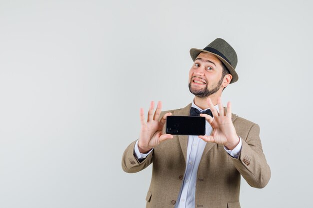 Hombre joven en traje, sombrero tomando fotos en el teléfono móvil y mirando alegre, vista frontal.