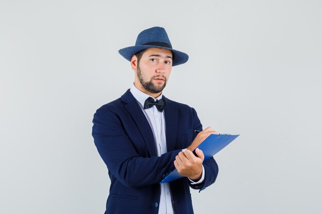 Hombre joven con traje, sombrero tomando notas en el portapapeles y mirando sensible, vista frontal.