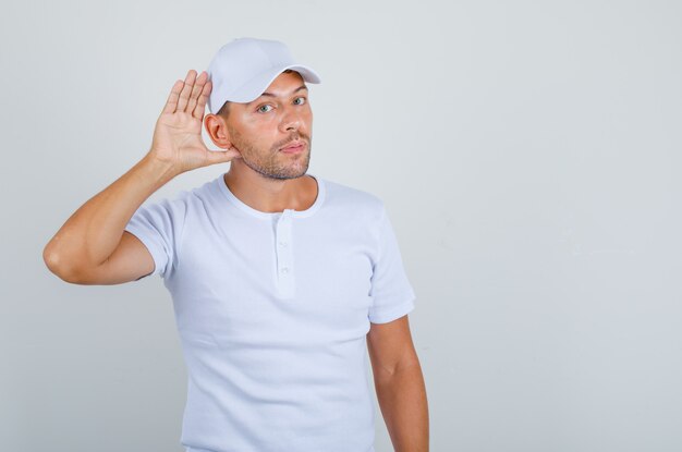 Hombre joven tratando de escuchar con gesto de oído en camiseta blanca, vista frontal de la tapa.