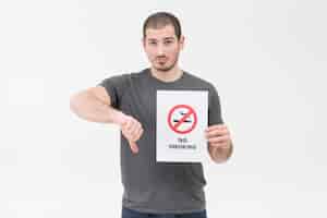 Foto gratuita el hombre joven triste que lleva a cabo la muestra de no fumadores que muestra el pulgar abajo gesticula contra el fondo blanco
