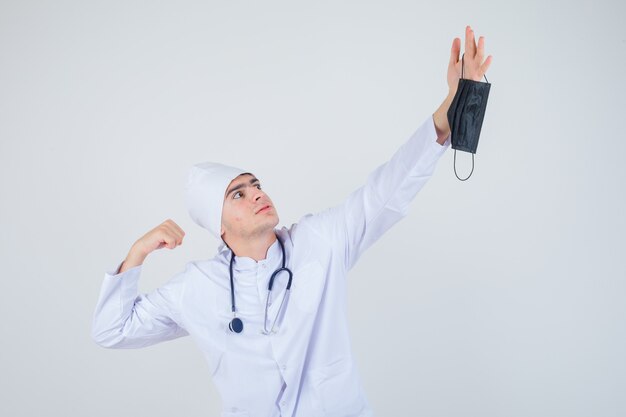 Hombre joven en uniforme blanco preparándose para llamar a la máscara médica y mirando enojado, vista frontal.