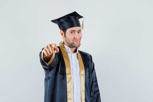 Foto gratuita hombre joven en uniforme graduado apuntando a la cámara y mirando confiado, vista frontal.