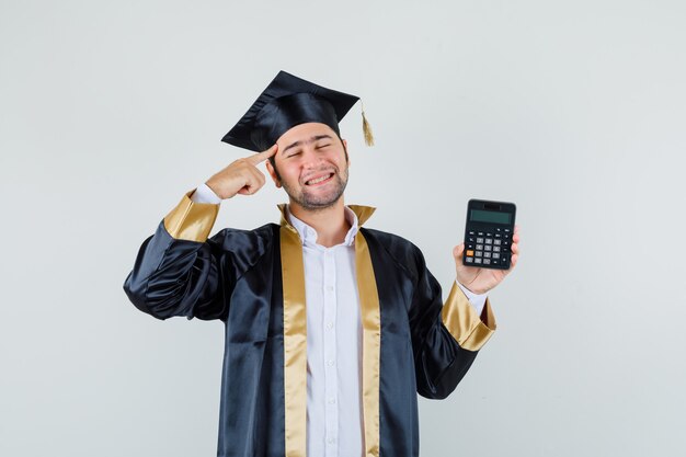 Hombre joven en uniforme graduado sosteniendo la calculadora con el dedo en las sienes y mirando contento, vista frontal.