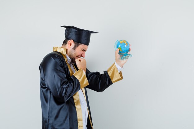 Hombre joven en uniforme graduado sosteniendo el globo de la escuela y mirando dichoso.