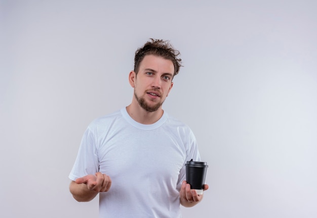 Hombre joven vestido con camiseta blanca sosteniendo una taza de café mostrando su gesto en la pared blanca aislada