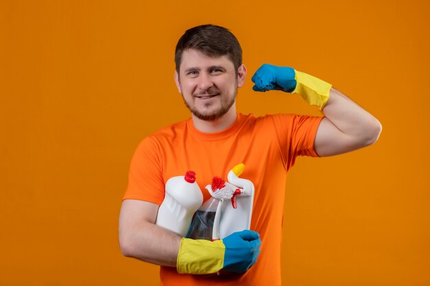 Hombre joven vestido con camiseta naranja y guantes de goma sosteniendo suministros de limpieza sonriendo alegremente positivo y feliz mirando a cámara mostrando bíceps listo para limpiar concepto sobre fondo naranja