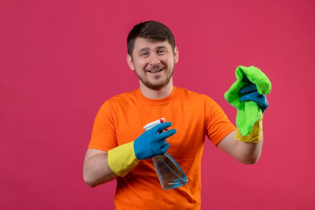 Hombre joven vestido con camiseta naranja y guantes de goma con spray de limpieza y alfombra sonriendo alegremente positivo y feliz de pie sobre la pared rosa