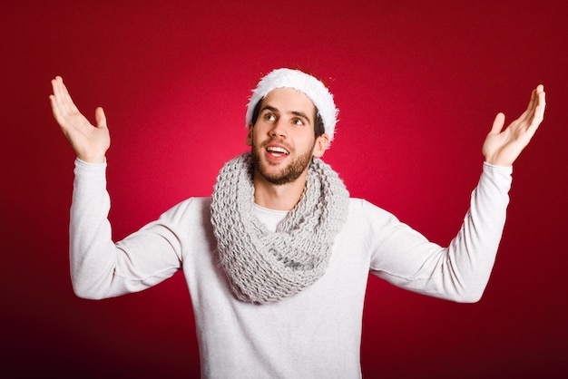 Hombre joven vestido con ropa de invierno bajo la nieve sobre fondo rojo chico con los brazos abiertos y sombrero de santa claus