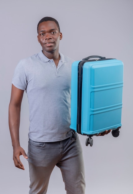 Hombre joven viajero afroamericano en camisa de polo gris con maleta azul positivo y feliz sonriendo amable