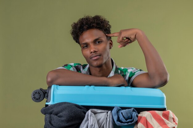 Hombre joven viajero afroamericano con maleta llena de ropa apuntando templo mirando confiado, centrado en la tarea sobre fondo verde