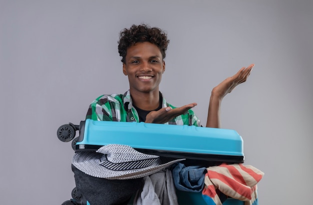 Hombre joven viajero afroamericano con maleta llena de ropa mirando a cámara sonriendo alegremente presentando con los brazos de sus manos sobre fondo blanco.