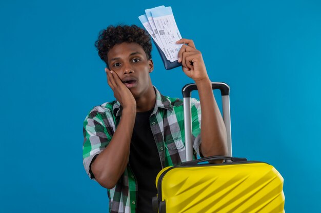 Hombre joven viajero afroamericano de pie con maleta sosteniendo billetes de avión mirando a la cámara sorprendido sobre fondo azul.
