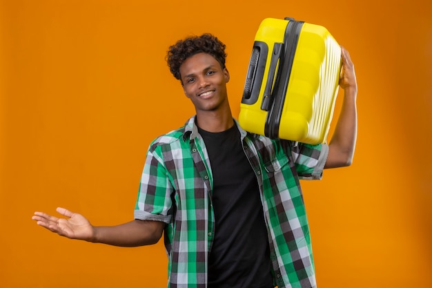 Hombre joven viajero afroamericano que sostiene la maleta mirando a la cámara sonriendo positivo y feliz extendiendo las manos haciendo gesto de bienvenida de pie sobre fondo naranja