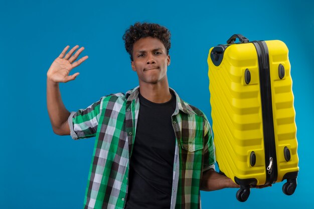 Hombre joven viajero afroamericano que sostiene la maleta saludando con la mano mirando a las cámaras con expresión escéptica en la cara de pie sobre fondo azul.