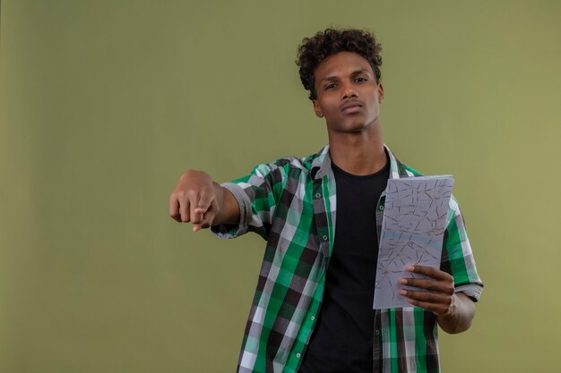 Hombre joven viajero afroamericano que sostiene el mapa apuntando a la cámara mirando a la cámara con expresión seria y confiada en la cara de pie sobre fondo verde