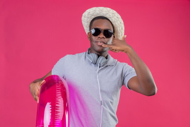 Hombre joven viajero afroamericano en sombrero de verano con auriculares alrededor de su cuello con gafas de sol negras sosteniendo anillo inflable sonriendo confiado haciendo gesto de llamarme sobre rosa