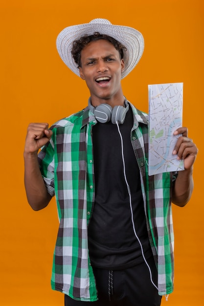 Hombre joven viajero afroamericano en sombrero de verano con auriculares sosteniendo mapa levantando puño salió y feliz levantando puño regocijándose de su éxito