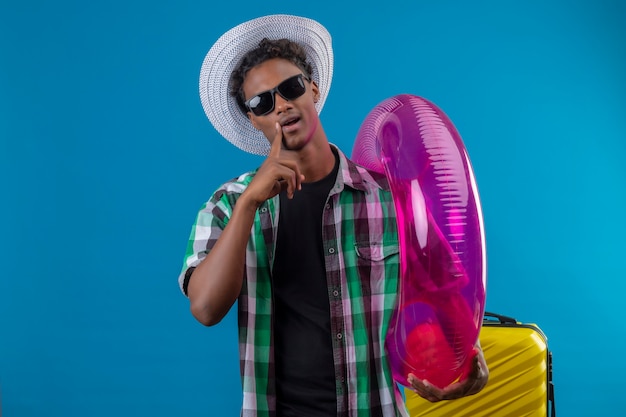 Hombre joven viajero afroamericano con sombrero de verano con gafas de sol negras con anillo inflable mirando a cámara con expresión de confianza sonriendo de pie sobre fondo azul
