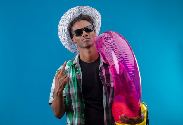 Hombre joven viajero afroamericano con sombrero de verano con gafas de sol negras sosteniendo un anillo inflable que parece seguro, satisfecho, mostrando el dedo medio