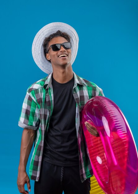 Hombre joven viajero afroamericano con sombrero de verano con gafas de sol negras sosteniendo anillo inflable sonriendo alegremente feliz y positivo de pie sobre fondo azul