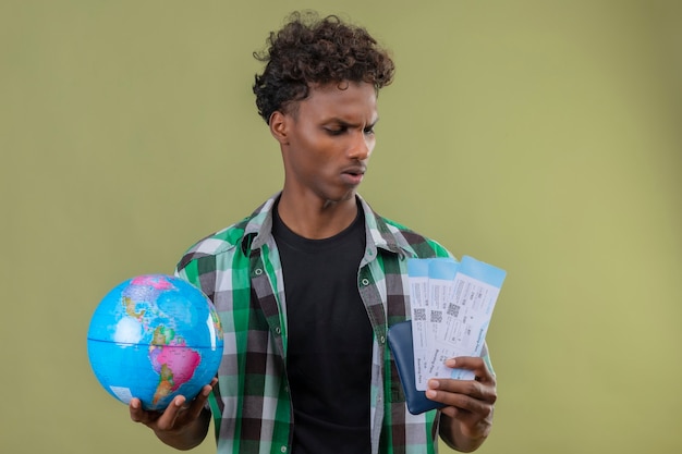 Hombre joven viajero afroamericano sosteniendo billetes de avión y globo terráqueo mirándolos con expresión seria en la cara, frunciendo el ceño