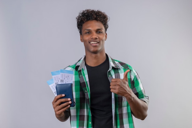 Hombre joven viajero afroamericano sosteniendo billetes de avión sonriendo alegremente positivo y feliz mirando a cámara mostrando los pulgares para arriba sobre fondo blanco