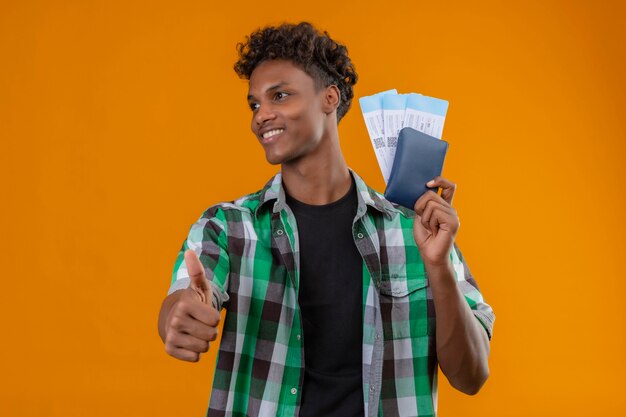 Hombre joven viajero afroamericano sosteniendo billetes de avión sonriendo alegremente positivo y feliz mirando a un lado mostrando los pulgares para arriba sobre fondo naranja
