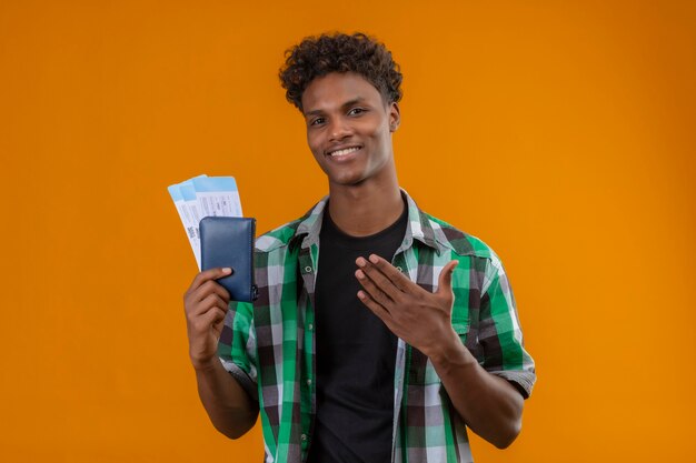 Hombre joven viajero afroamericano sosteniendo billetes de avión sonriendo alegremente positivo y feliz presentando con el brazo de su mano mirando a la cámara de pie sobre fondo naranja