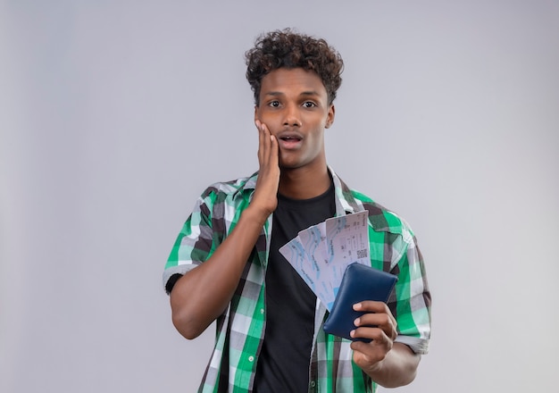 Hombre joven viajero afroamericano sosteniendo billetes de avión sorprendido y asombrado mirando a la cámara que cubre la polilla con la mano de pie sobre fondo blanco.