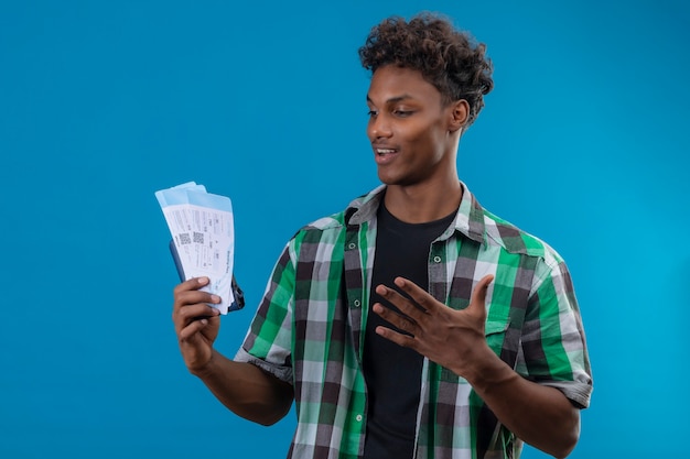Hombre joven viajero afroamericano sosteniendo boletos de avión sonriendo alegremente positivo y feliz mirando boletos salió de pie sobre fondo azul