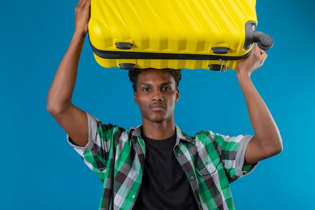 Hombre joven viajero afroamericano sosteniendo la maleta en la cabeza mirando a la cámara con expresión seria de confianza en la cara de pie sobre fondo azul