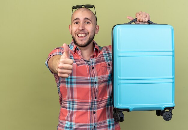 Hombre joven viajero alegre que sostiene la maleta y pulgar hacia arriba aislado en la pared verde oliva con espacio de copia
