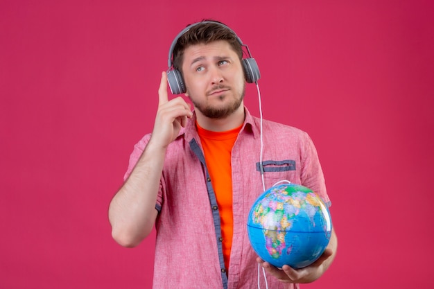 Hombre joven viajero con auriculares sosteniendo globo escuchando música sonriendo de pie positivo sobre fondo rosa