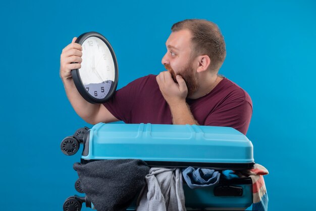 Hombre joven viajero barbudo con maleta llena de ropa sosteniendo el reloj de pared mirándolo en pánico