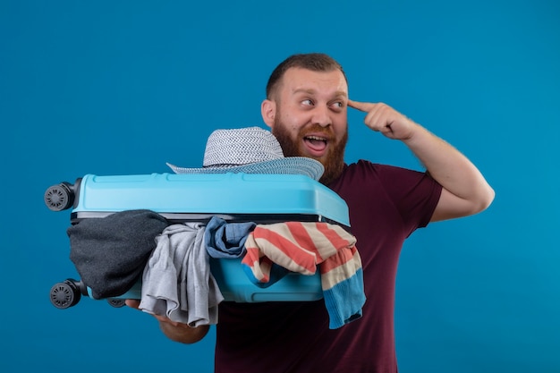 Hombre joven viajero barbudo que sostiene la maleta llena de ropa mirando a un lado apuntando templo teniendo nueva idea