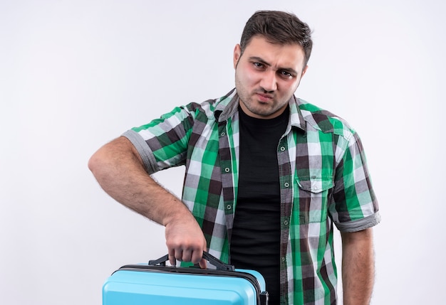 Foto gratuita hombre joven viajero en camisa a cuadros con maleta con expresión triste en la cara de pie sobre la pared blanca