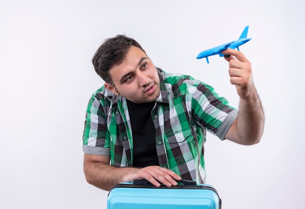 Hombre joven viajero en camisa a cuadros de pie con maleta sosteniendo avión de juguete sonriendo con cara feliz sobre pared blanca