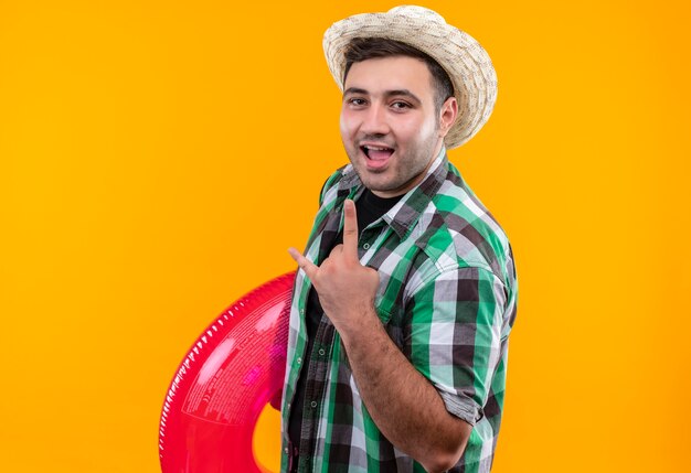 Hombre joven viajero en camisa a cuadros y sombrero de verano con anillo inflable feliz y salido mostrando el símbolo de la roca con los dedos de pie sobre la pared naranja