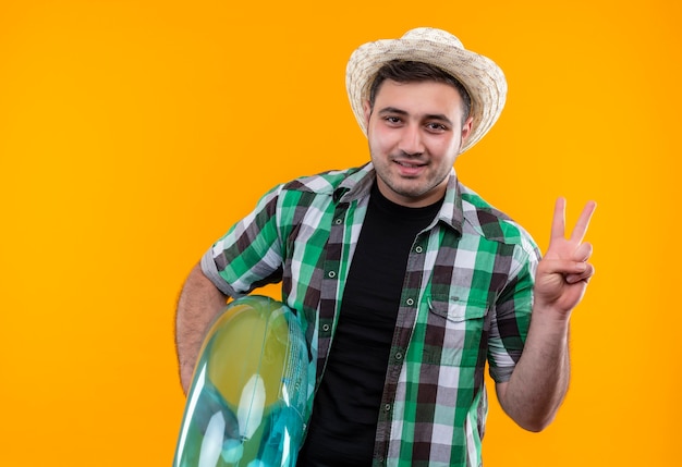 Hombre joven viajero en camisa a cuadros y sombrero de verano con anillo inflable que muestra el signo de la victoria sonriendo de pie sobre la pared naranja