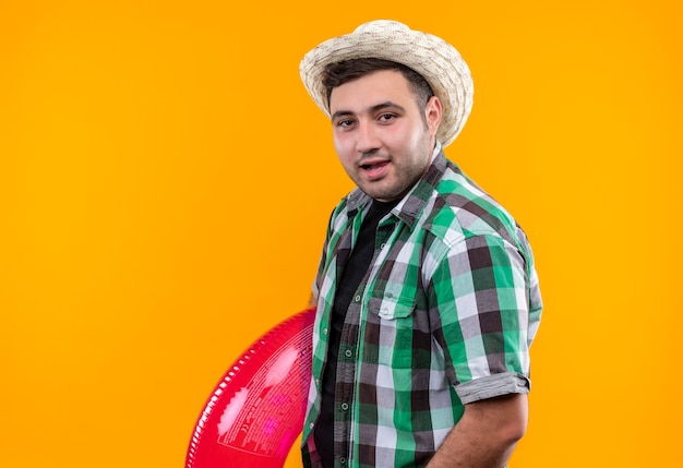 Hombre joven viajero en camisa a cuadros y sombrero de verano con anillo inflable con una sonrisa en la cara de pie sobre la pared naranja