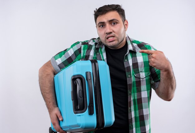 Hombre joven viajero en camisa a cuadros sosteniendo la maleta confundido apuntando con el dedo a sí mismo de pie sobre la pared blanca