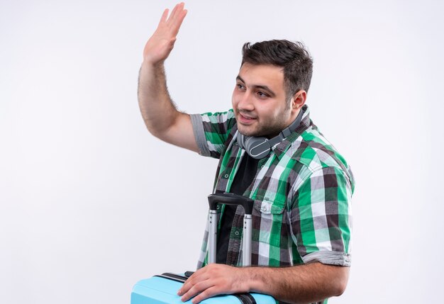 Hombre joven viajero en camisa a cuadros sosteniendo la maleta mirando a un lado sonriendo saludando con la mano de pie sobre la pared blanca