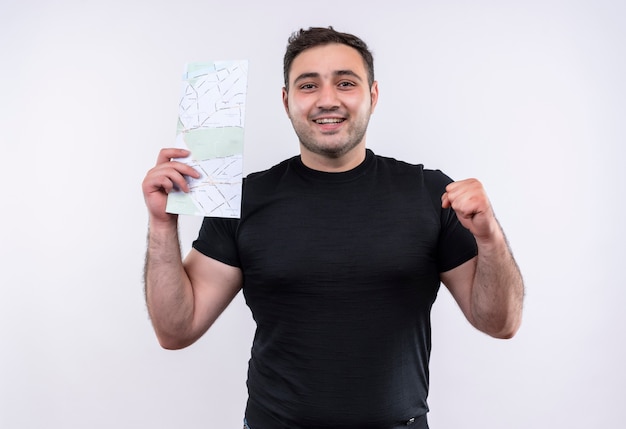 Hombre joven viajero en camiseta negra con billetes de avión feliz y positivo puño sonriendo de pie sobre la pared blanca