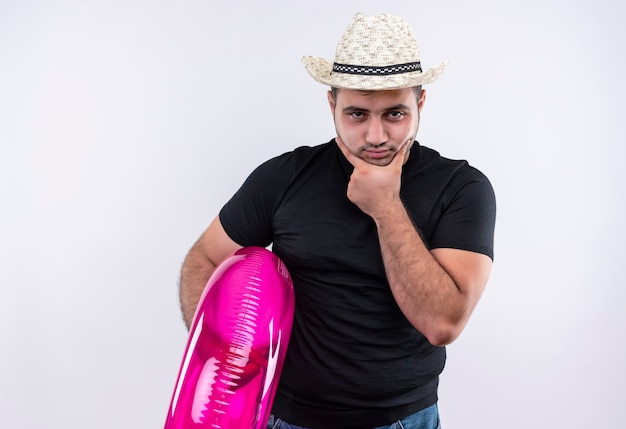 Foto gratuita hombre joven viajero en camiseta negra y sombrero de verano sosteniendo un anillo inflable con el ceño fruncido y perplejo parado sobre la pared blanca