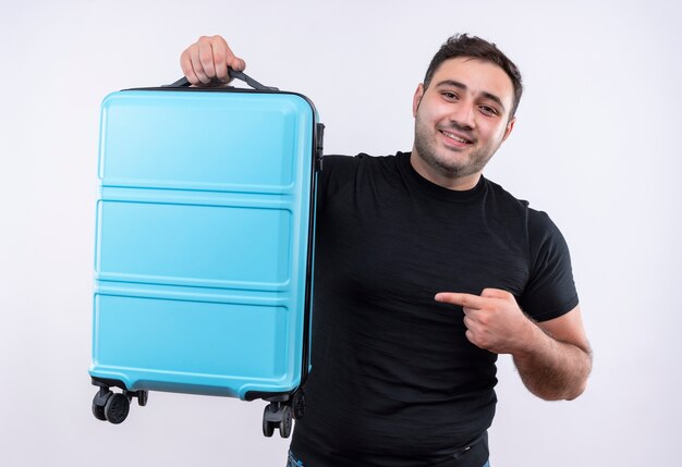 Hombre joven viajero en camiseta negra sosteniendo la maleta apuntando con el dedo a ella sonriendo confiado de pie sobre la pared blanca