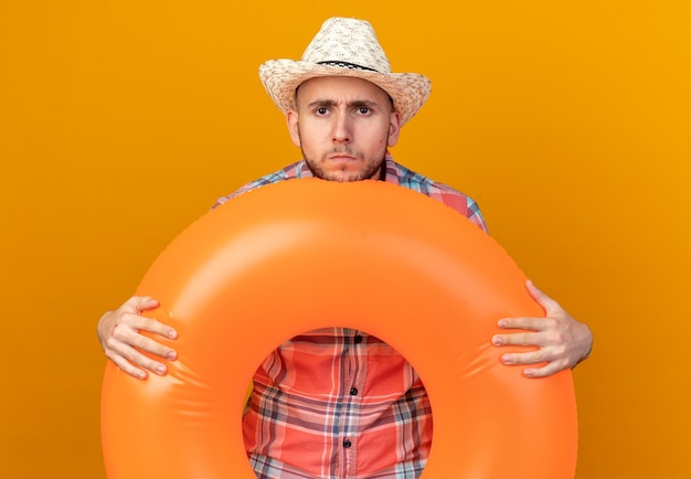Foto gratuita hombre joven viajero descontento con sombrero de playa de paja sosteniendo el anillo de natación aislado en la pared naranja con espacio de copia