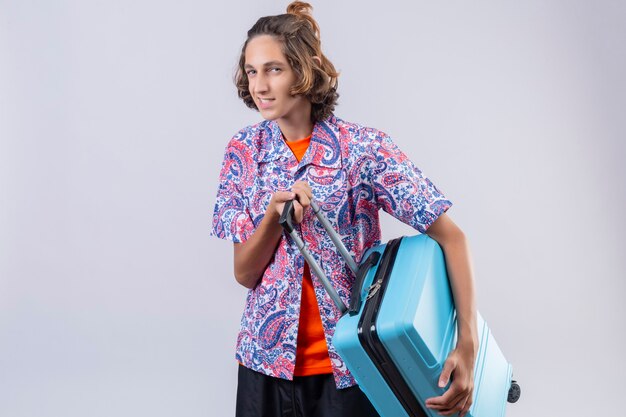 Hombre joven viajero con maleta azul mirando confiado sonriendo con cara feliz listo para viajar de pie sobre fondo blanco.