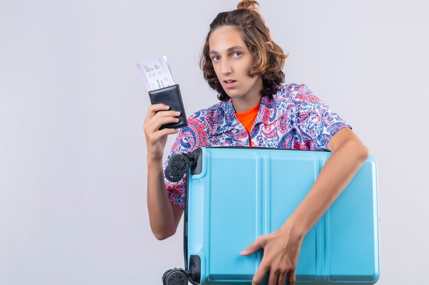 Hombre joven viajero con maleta sosteniendo billetes de avión mirando confundido parado sobre fondo blanco.