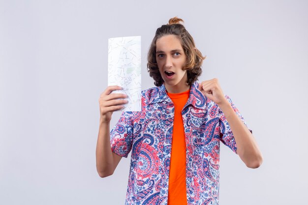 Hombre joven viajero con mapa levantando el puño después de una victoria feliz y salido de pie sobre fondo blanco.