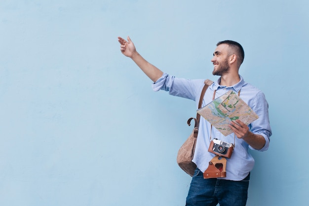 Hombre joven viajero mostrando algo con la celebración de mapa de pie contra el fondo azul.